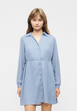 Ung kvinde iført en lyseblå, langærmet, knap-op kjole med taljeindsnøring og diskret prikket mønster, stående foran en ensfarvet baggrund.