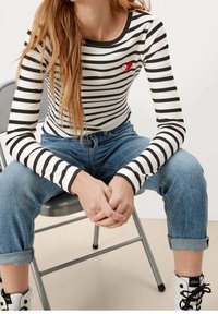 Czarnobiała prążkowana bluzka z długim rękawem, okrągłym dekoltem i małym czerwonym serduszkiem, zestawiona z podwiniętymi niebieskimi dżinsami i białymi botkami.