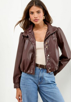 Vrouw met lang bruin haar, draagt gouden oorbellen, een bruine leren knopenjack, een beige top, blauwe jeans en een bruine riem.