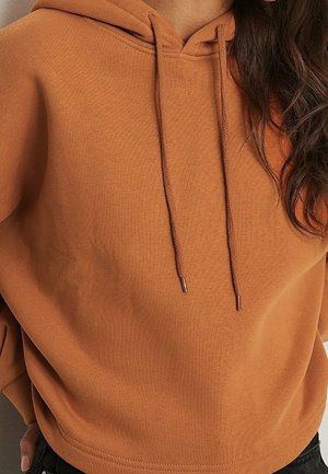 Kvinde iført en brun hættetrøje med snore og sorte bukser, der viser overkroppen og bølget mørkt hår over den ene skulder.