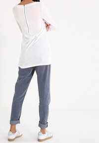 Top bianco a maniche lunghe con zip sul retro, abbinato a pantaloni leggeri blu chiaro larghi e arrotolati, e scarpe bianche con dettagli verdi.