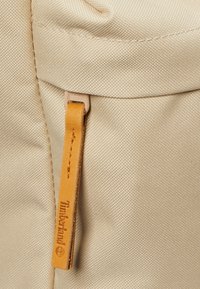 Zaino in tessuto beige con superficie testurizzata, dotato di una linguetta zip in pelle color marrone chiaro con logo Timberland inciso e dettagli di cucitura.