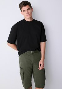 Young man standing against white wall wearing black oversized t-shirt and olive green cargo shorts with belt, looking at camera.