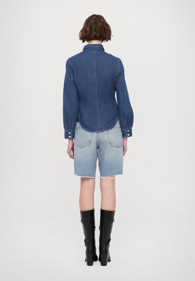 Chemise en denim bleu foncé à manches longues, avec des poignets boutonnés et un ourlet arrondi, associée à un short en jean clair effet usé et des bottes noires jusqu'au genou.