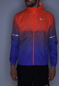 Chaqueta cortavientos Nike con un degradado de naranja a morado, detalles reflectantes plateados, cremallera frontal y capucha. Tejido suave y ligero.