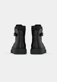 Botas de cuero negro hasta el tobillo con un acabado texturizado, que presentan un cuello acolchado y un detalle de hebilla lateral, montadas sobre una sólida suela de goma negra.