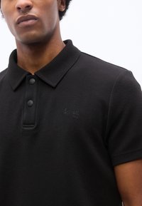Homme portant un polo noir texturé avec une patte de boutonnage et un logo embossé "Schott NYC" sur le côté gauche de la poitrine.
