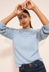 Svetlomodrý pletený sveter s okrúhlym výstrihom a textúrovaným vzorom na ramenách, doplnený tmavými džínsami. Mäkký, pohodlný materiál, rebrované manžety.