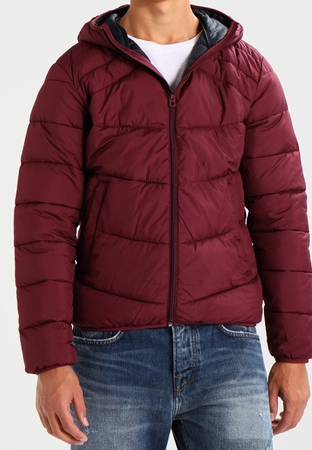 Man som bär en zipad vinröd vadderad jacka med huva och två sidofickor över en vit t-shirt och blå jeans.
