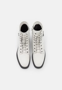 Bottes de cheville en cuir blanc avec semelles noires, dotées d'un laçage à l'avant et d'une fermeture éclair. Texture lisse avec une forme structurée et des accents en métal argenté.