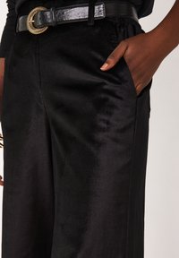Pantalon noir en satin avec une texture lisse, doté d'une poche latérale et d'une ceinture noire brillante avec une boucle décorative en or.