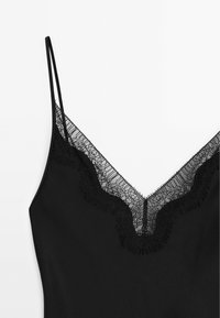 Camisola nera con spalline sottili, scollo a V caratterizzato da un intricato bordo in pizzo nero e bordi frangiati, texture del tessuto liscia.