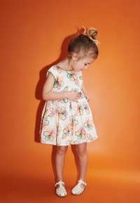 Weißes Blumenkleid mit buntem Stickerei, kurzen Ärmeln und einem schwingenden Rock. Kombiniert mit weißen Sandalen an einem Kind vor einem orangefarbenen Hintergrund.