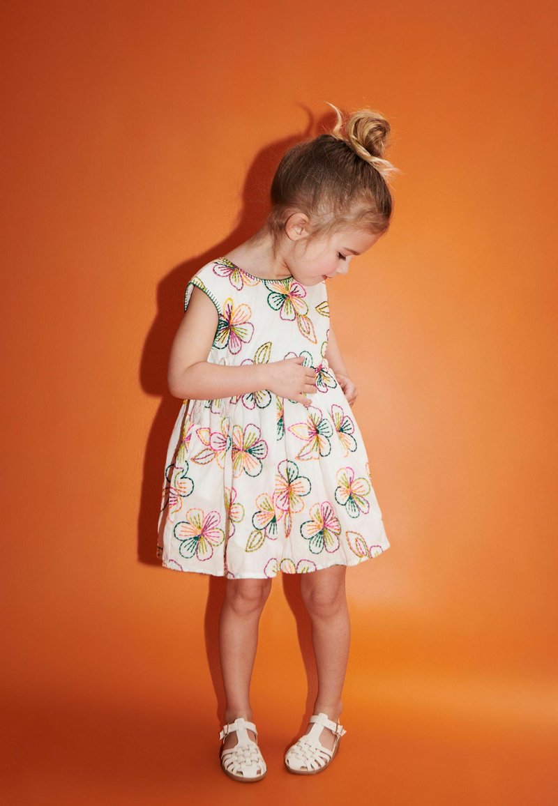 Weißes Blumenkleid mit buntem Stickerei, kurzen Ärmeln und einem schwingenden Rock. Kombiniert mit weißen Sandalen an einem Kind vor einem orangefarbenen Hintergrund.