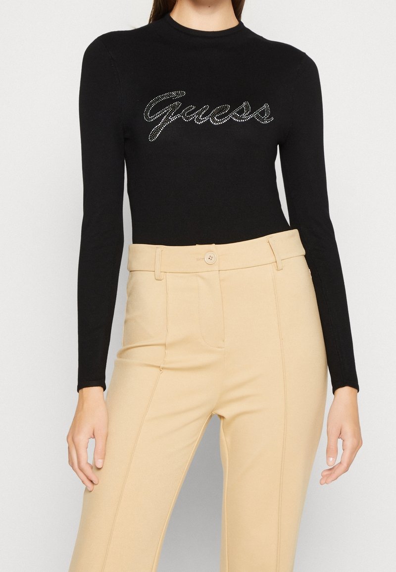 Schwarzer Langarmbodysuit mit rundem Ausschnitt, versehen mit einem funkelnden "Guess"-Logo aus silbernen Strasssteinen; kombiniert mit beigen, hochgeschnittenen Hosen.