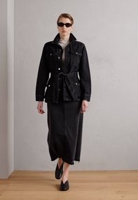 Černá džínová bunda se zavazováním v pase, doplněná dlouhou černou džínovou sukní. Sluneční brýle a krémový top s volánky dokončují outfit.