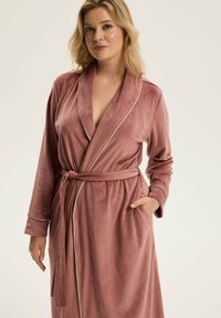 Różowy aksamitny szlafrok z długimi rękawami, kołnierzem szalowym i paskiem do przewiązywania. Posiada boczne kieszenie oraz delikatne białe wykończenie wzdłuż brzegów.