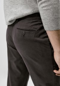 Personne portant un pantalon marron foncé avec une poche arrière à bouton et un pull gris clair, se tenant avec la main détendue à ses côtés.