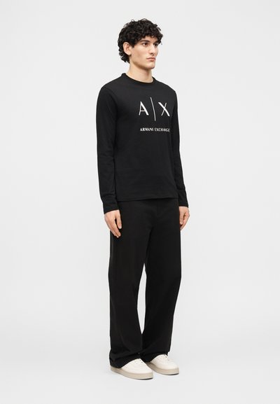 Млад мъж, облечен с черна риза с дълги ръкави Armani Exchange, черен панталон и бели маратонки, изправен пред едноцветен фон.