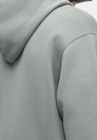 Gros plan d'une personne portant un sweat à capuche gris clair, montrant les détails du dos et de la couture d'épaule du vêtement.