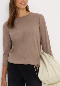 Femme portant un haut à manches longues marron clair avec un détail de noeud sur le côté, un pantalon noir, tenant un grand sac en tissu doux crème.