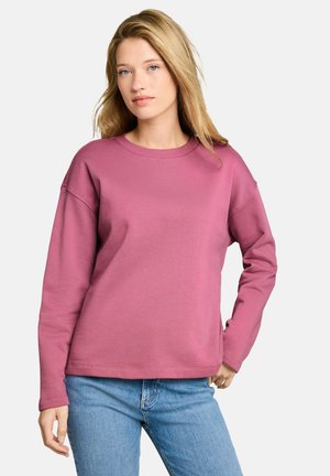Frau mit blondem Haar, die einen langärmligen pinken Pullover und blaue Jeans trägt, steht mit einer Hand in der Hüfte.
