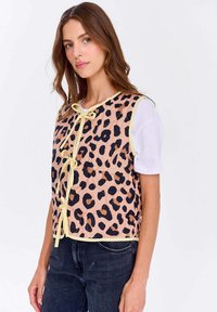 Žena s dlouhými hnědými vlasy, která má na sobě bezrukávovou vestu s leopardím potiskem a žlutým lemem přes bílé tričko s krátkým rukávem a tmavé džíny.