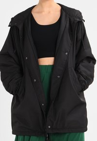 Chaqueta negra, ligera, con capucha, que presenta un cierre de botones en el frente, puños elásticos y dos bolsillos laterales, llevada sobre una camiseta negra y pantalones verdes.