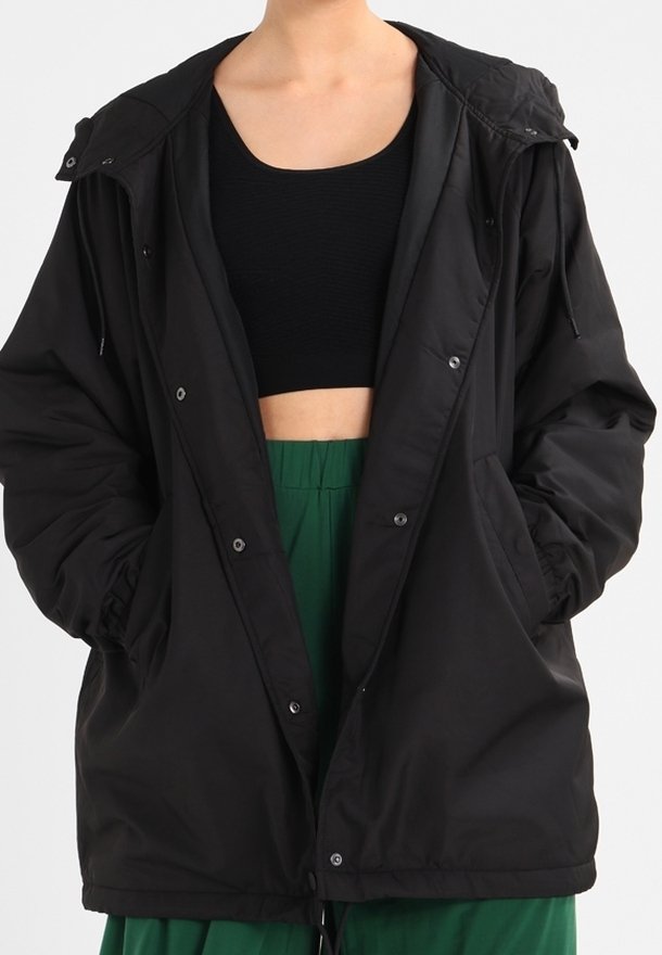 Chaqueta negra, ligera, con capucha, que presenta un cierre de botones en el frente, puños elásticos y dos bolsillos laterales, llevada sobre una camiseta negra y pantalones verdes.