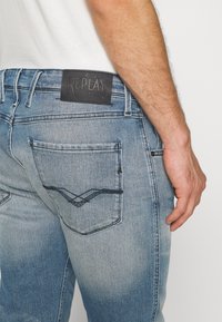 Jean en denim bleu clair avec des détails délavés, coutures visibles sur la poche arrière et un patch en cuir noir avec la marque "Replay" près de la taille.