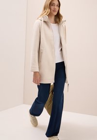 Cappotto in pile beige chiaro con cappuccio, dotato di chiusura con zip e tasche laterali. Abbinato a pantaloni blu scuro e sneakers beige.