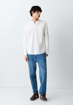 Giovane uomo in piedi, indossa una maglia henley bianca a maniche lunghe, jeans blu e sneakers marroni, guarda verso sinistra.