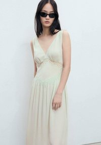 Long, pale green dress with a deep V-neck, lace accents at the waist, and flowing skirt. Model wearing black sunglasses, standing against a light background.