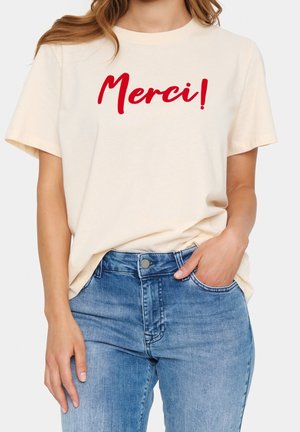Kobieta w kremowej koszulce z czerwonym tekstem "Merci!" i niebieskich dżinsach, ręka w kieszeni, widok z przodu przycięty na wysokości szyi.