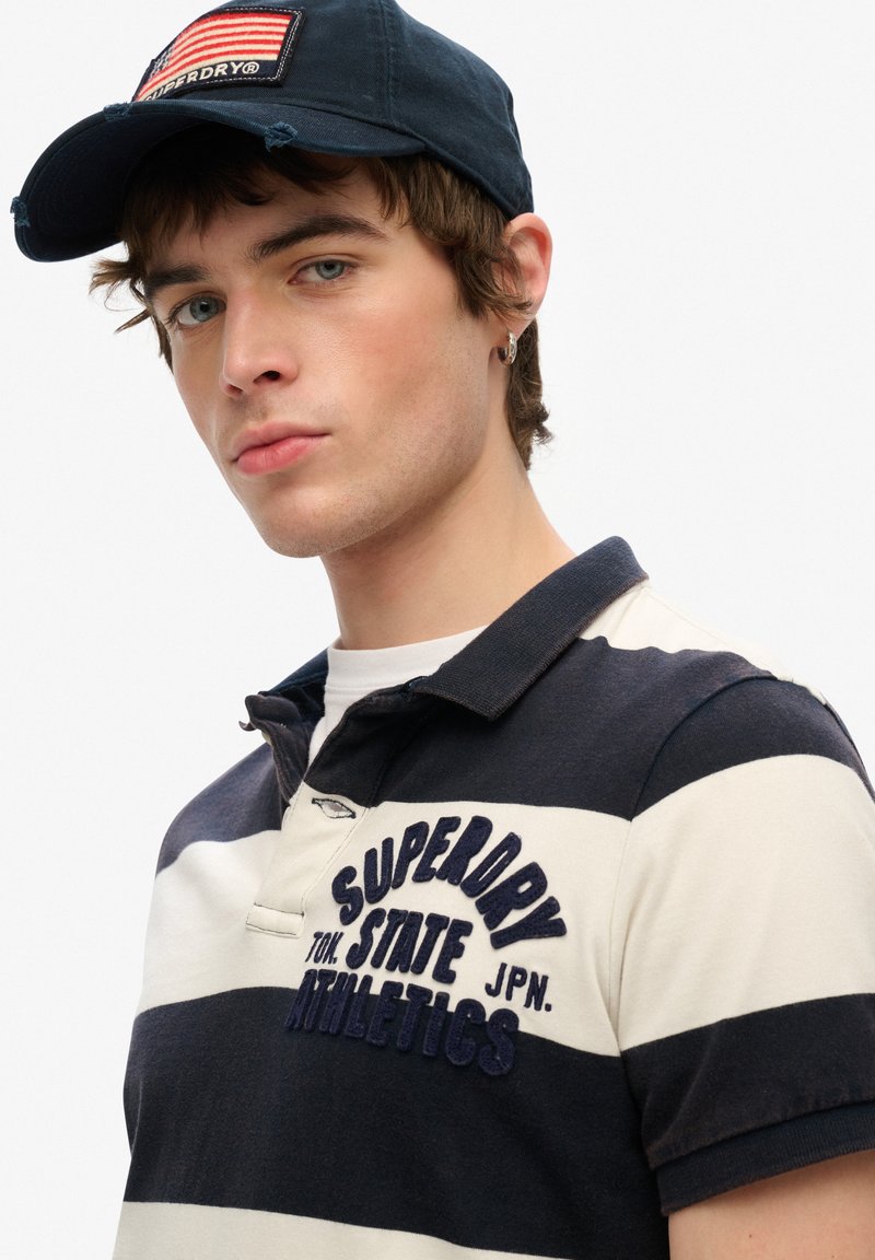 Camiseta polo a rayas en azul marino y crema con el logotipo bordado de "Superdry". Combinada con una gorra azul marino con un parche de bandera. Corte casual.