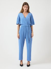 Kvinne står iført en lys blå, belagt jumpsuit med plisseringer og vide ermer, samt svarte spisse hæler, mot en enkel hvit bakgrunn.