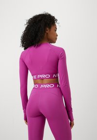 Ljubičasti dugih rukava atletski top s krojenjem koje otkriva struk, s širokim "Nike Pro" logom na dnu i glatkom teksturom.