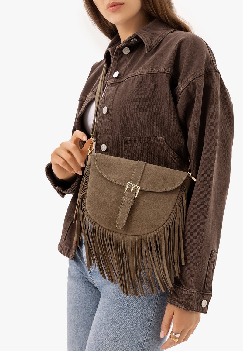 Femme portant une veste marron boutonnée et un jean, tenant un sac bandoulière en suède marron à franges avec fermeture à boucle.
