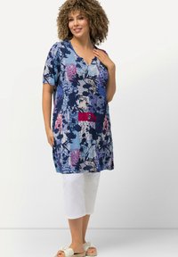 Modrá a fialová tunika se vzorem, krátkými rukávy, výstřihem do V a bočními rozparky, v kombinaci s bílými zkrácenými kalhotami a světlými sandály.