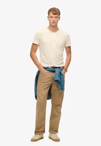 Beige kortærmet henley skjorte, tan chinos, denimskjorte bundet om taljen, beige sneakers med strukturerede såler, enkelt design.