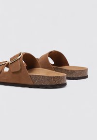 Sandales en daim marron avec deux larges sangles, perforations circulaires et une semelle en liège texturée. Comprend un accent en boucle métallique sur une des sangles.