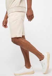 Shorts en denim beige avec des revers, présentés sur une jambe. Associés à des baskets blanches avec un design texturé et une semelle claire.