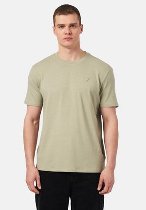 Młody mężczyzna z krótkimi brązowymi włosami, ubrany w jednolity jasnobeżowy krótki rękaw t-shirt i czarne spodnie, stojący na białym tle.