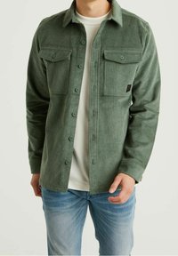 Vihreä pikeepaita, jossa napillinen etuosa, kaksi rintataskua ja poimuttainen pinta. Tämä on puettu valkoisen T-paidan päälle ja yhdistetty vaaleansinisiin farkkuihin.