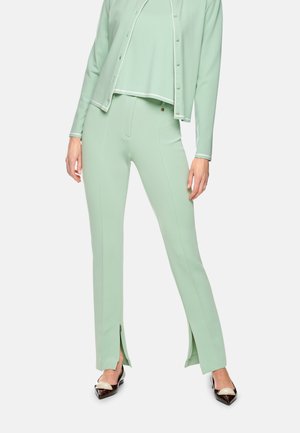 Woman wearing light green high-waist pants with front ankle slits, matching buttoned cardigan, and black and beige pointed flats.