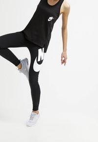 Černý tílko s logem Nike, spárované s úzkými černými legínami s velkým bílým logem Nike. Šedé sportovní boty dotvářejí celé oblečení.