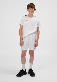 Weißes Sporttrikot und Shorts mit schwarzen Akzenten und Logo, kombiniert mit schwarzen Sportschuhen und weißen Socken. Der Stoff wirkt leicht.