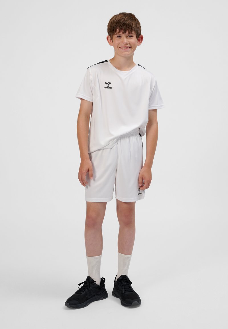 Weißes Sporttrikot und Shorts mit schwarzen Akzenten und Logo, kombiniert mit schwarzen Sportschuhen und weißen Socken. Der Stoff wirkt leicht.