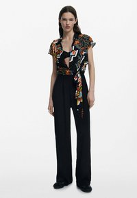 Black jumpsuit with a short-sleeved, patterned, tied front top. The design features floral and geometric prints, creating a bold contrast.