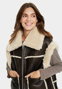 Ruskea shearling-vuori liivi, jossa on pehmeä beige kaulus, vetoketjuluukku ja etutaskut. Ominaisuuksiin kuuluu kontrastivalkoinen ommel saumoissa.